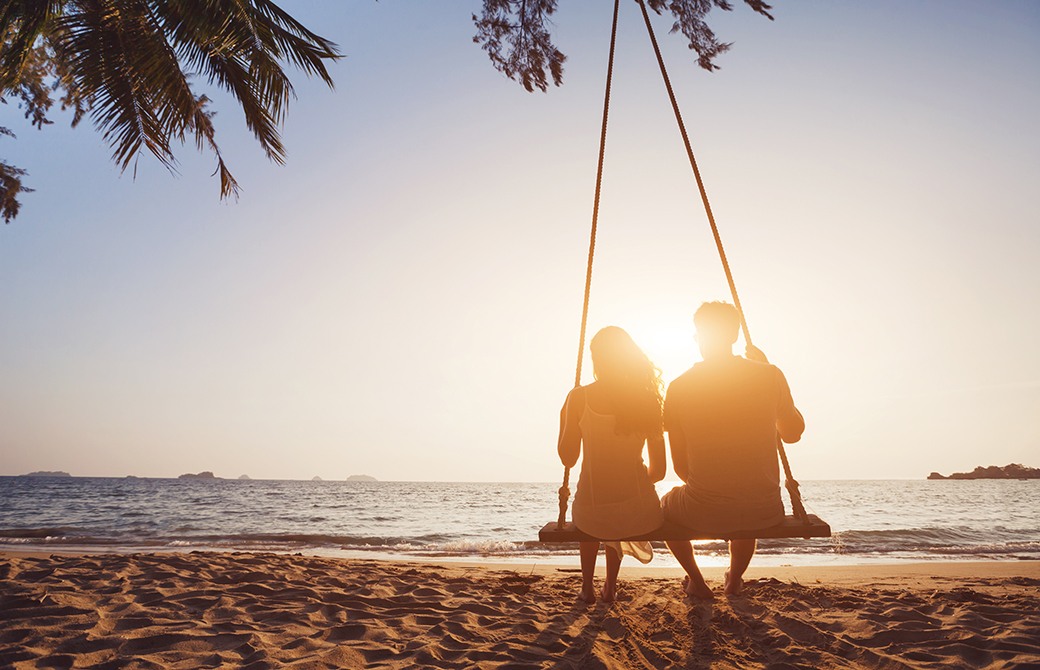 夕暮れ時の海辺の砂浜で肩を寄せ合ってブランコに乗る男女