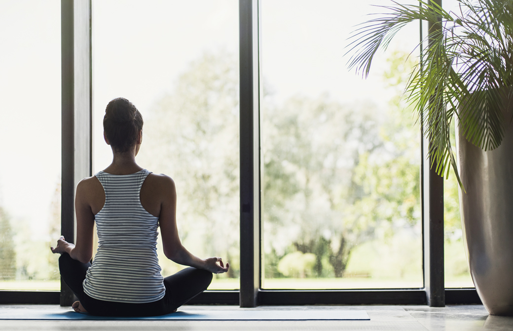 室内の窓際でヨガマットの上にあぐらで座りこちらに背を向けて瞑想をしている女性
