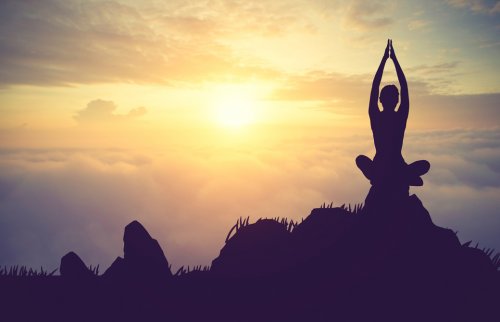 小高い岩の上で朝日に向ってあぐらで座り腕を頭上に伸ばして合掌している女性