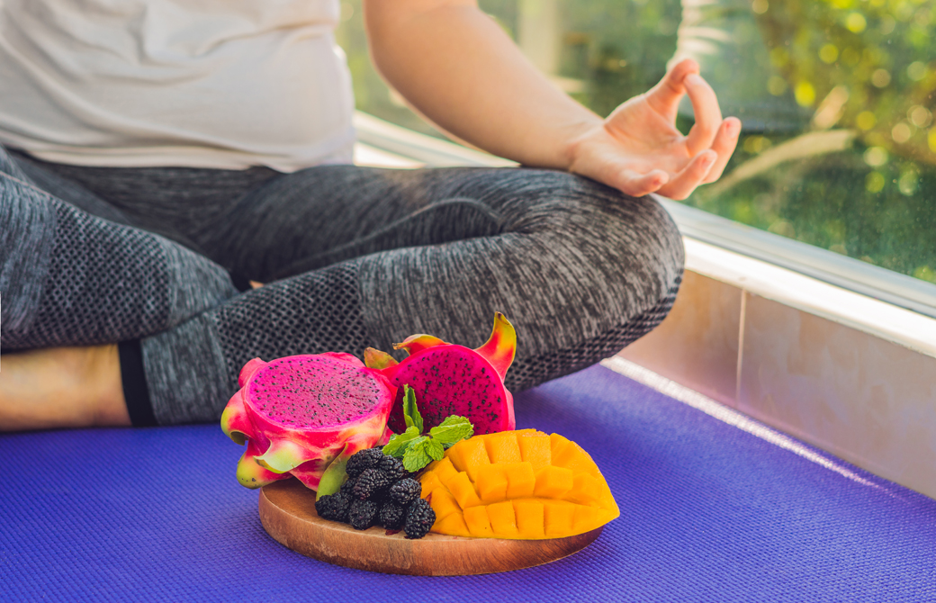 ヨガマットの上でフルーツの盛られた器を前にあぐらの姿勢をとる女性