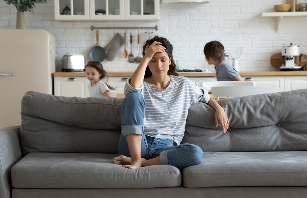 ソファの上でおでこを抱えて疲れている母親と後ろで走り回る幼い兄妹