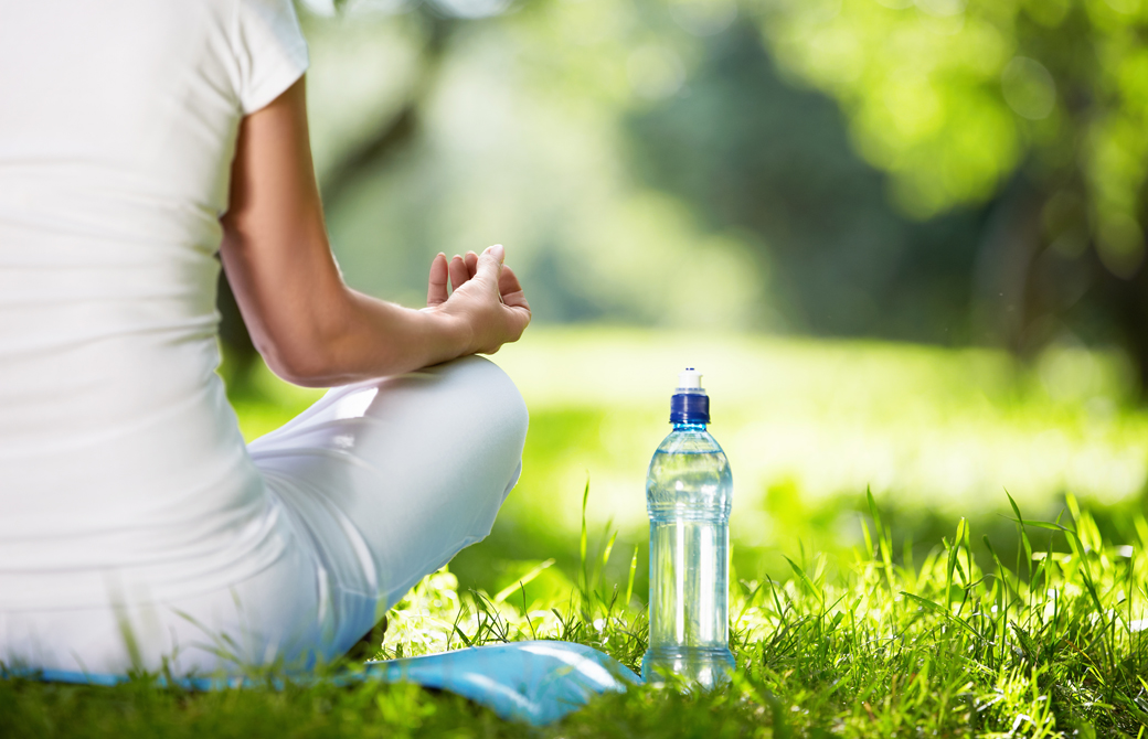 木漏れ日のある公園の芝生であぐらをして水のボトルを横に置いている女性