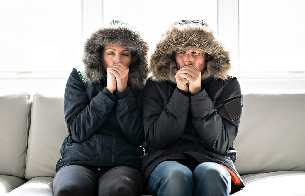 ソファでダウンジャケットを着た男女が寒そうに手をこすり合わせている