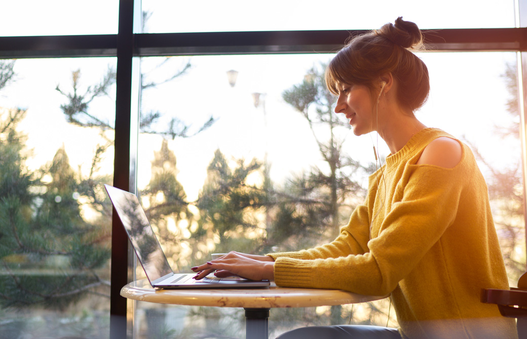 窓際で微笑みながらPCに向ってタイピングしている黄色いニットを着た女性