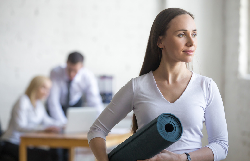 オフィスでヨガマットをもっている女性と背後にいるビジネスマン風の男性たち