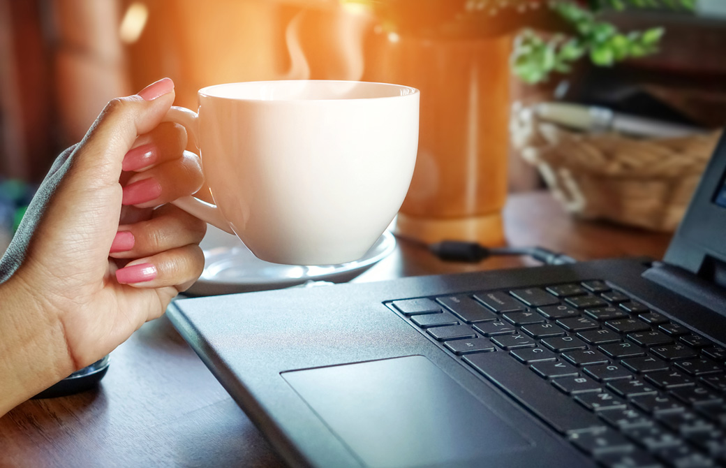 PCの前で湯気の出るコーヒーカップを持っている女性の手元