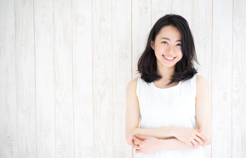 白い壁を背にして白いワンピースの女性が肘を組んで微笑んでいるウエストアップの写真