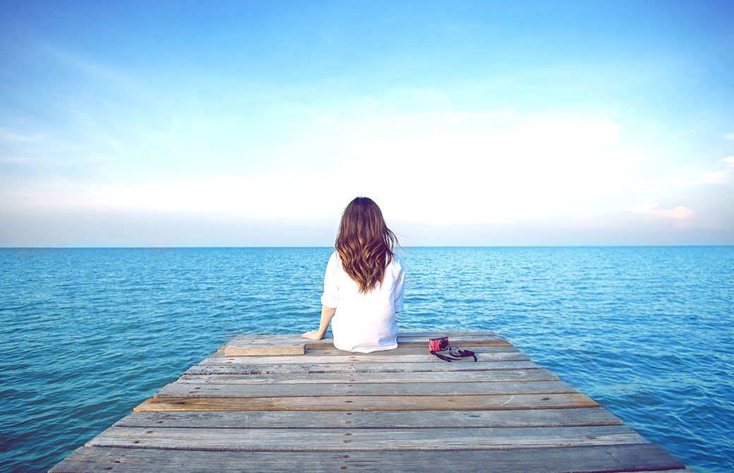 青空と海が広がる中、女性が1人で後ろ向きに座っている写真