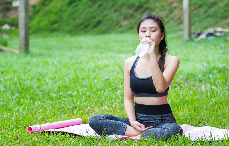 芝生の上にヨガマットを敷いて水を飲んでいる女性