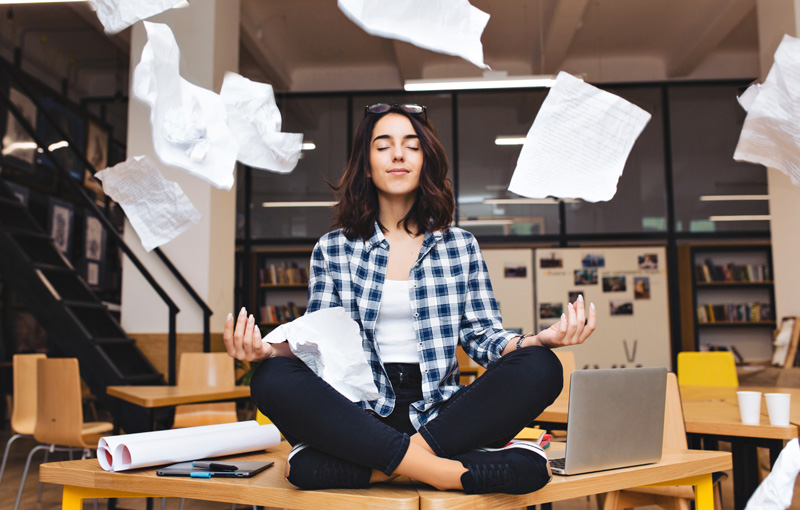 紙がバラまかれている中で目を閉じてあぐらを組んだ女性が机に座っている