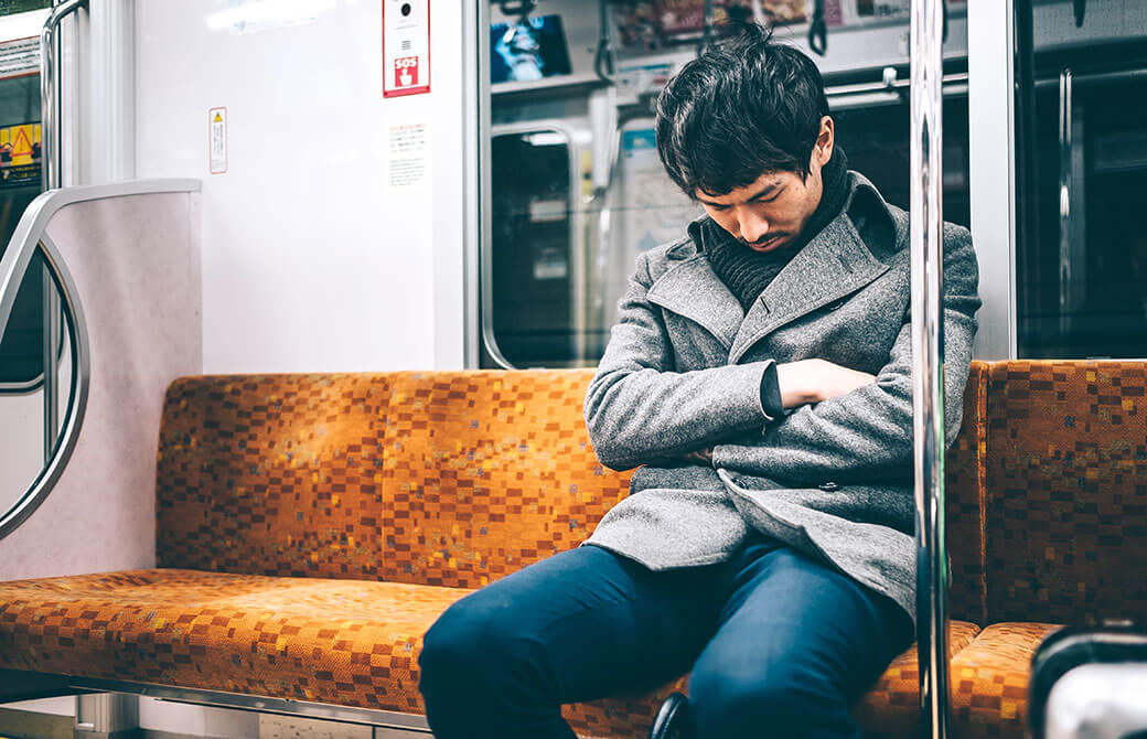 電車の中で居眠りするコートを羽織った男性の写真