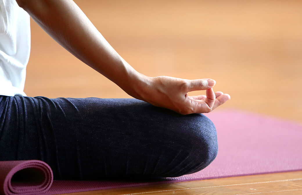 床の上に敷いたピンクの待ったの上であぐらをかいて瞑想をしている人の脚と手の写真