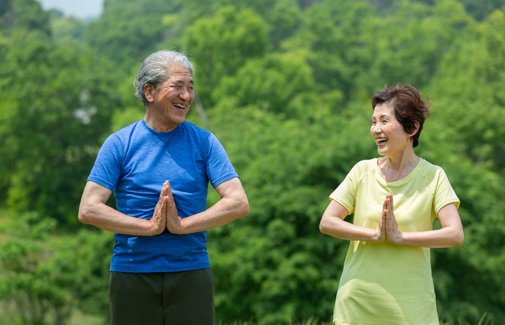 緑の木の前で高齢の男性と女性が合掌をして、目を合わせて微笑んでいる