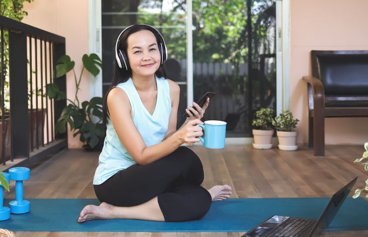 スマホとマグカップを手にヘッドフォンでヨガを楽しむ女性
