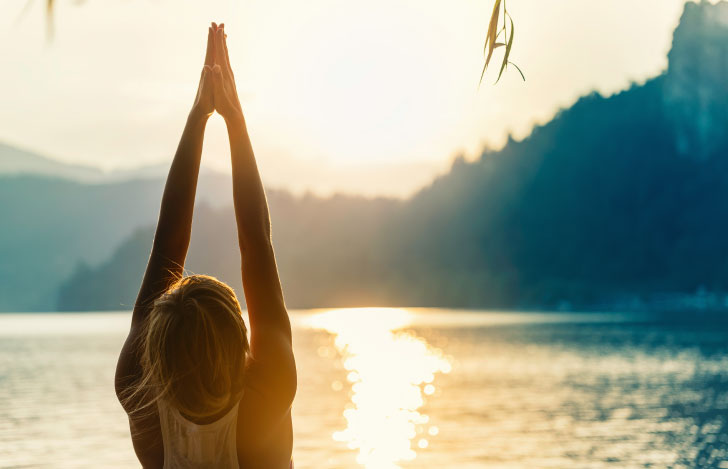 太陽に向かって両腕を上げて手を合わせる女性の後ろ姿