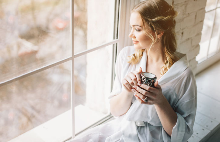 白い着物を羽織り窓辺でカップを手に外を眺める女性