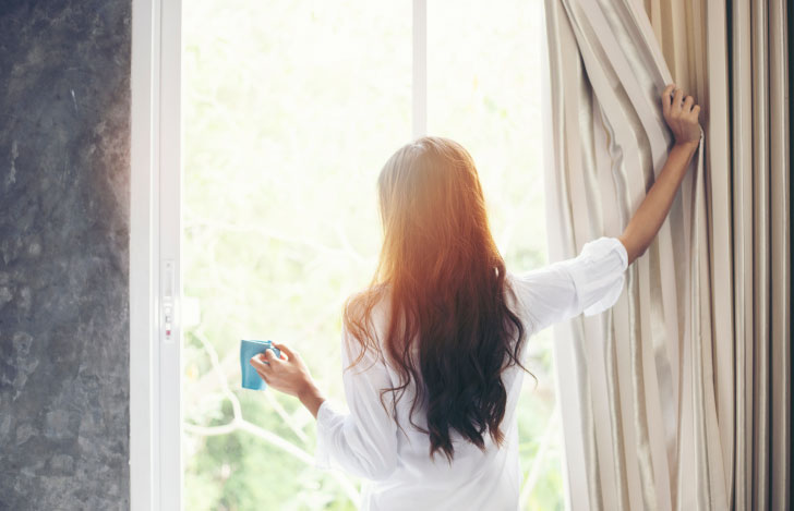 カーテンを開けて朝日を浴びる女性の後ろ姿