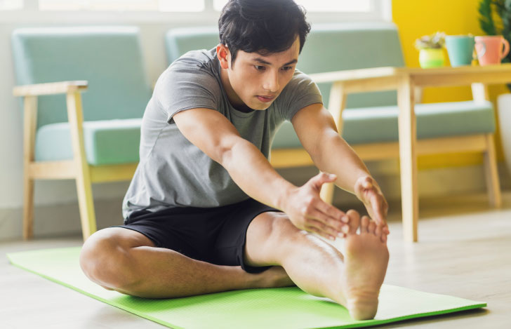 ヨガマットの上に座り、片脚を伸ばして前屈をする男性