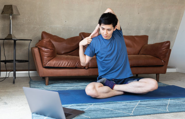 自室のヨガマットの上でPC画面を見ながらヨガを行う男性
