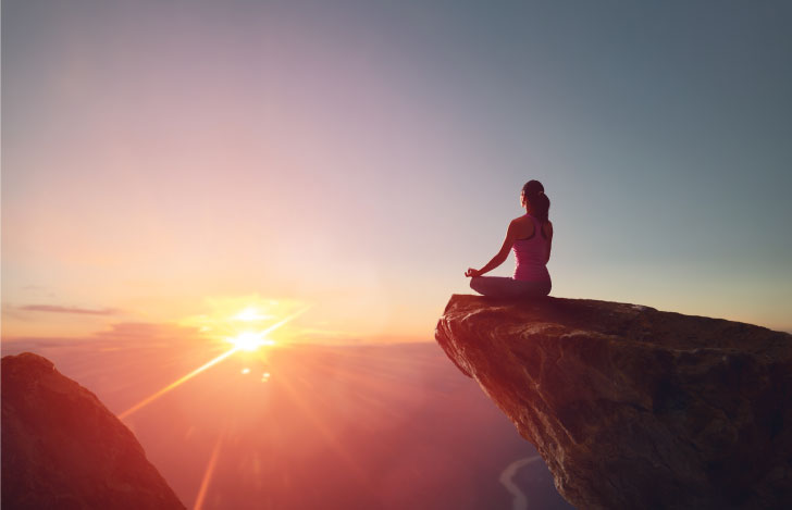 せり出した岩地の先端で夕日に向かって瞑想する女性