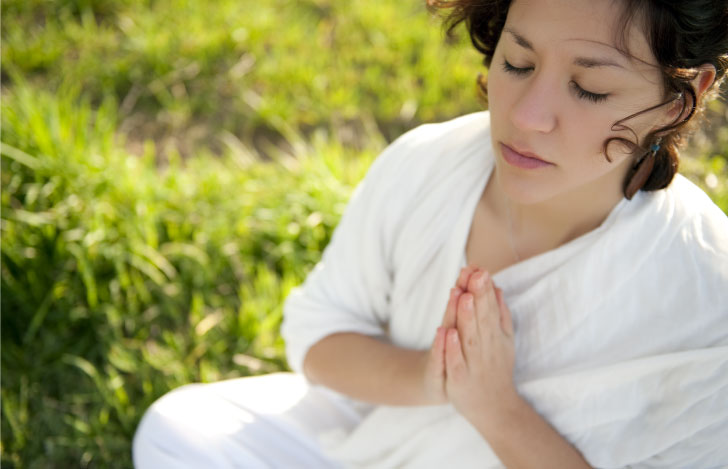 草原に座り胸の前で手を合わせて目を閉じる女性