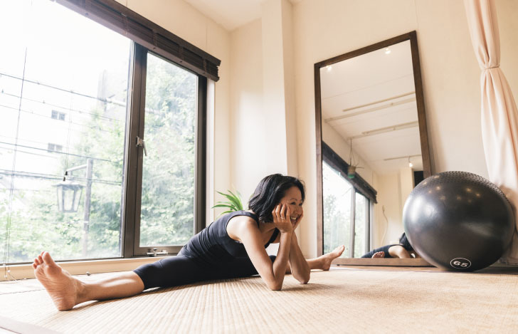 床の上で開脚をしながら頬杖をつく笑顔の女性