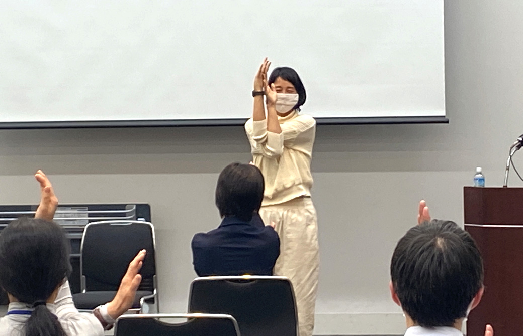 前で立ってポーズを取るいずみ先生と、スーツ姿で座ってポーズを取る参加者