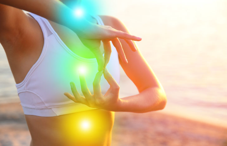 海を背景にヨガのポーズをとる女性の手元とチャクラを表す光