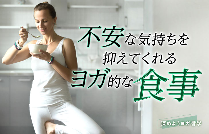 キッチンで立ち木のポーズをとりながらボウルを抱えて食事する白いヨガウェアの女性