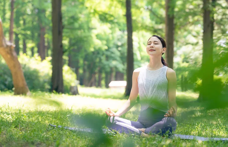 木々が茂る自然の中で目を閉じ心地良さそうに呼吸するヨガウェアを着た女性