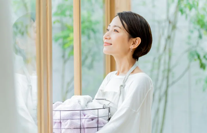 洗濯物のかごを抱えながら笑顔で窓の外を眺める女性