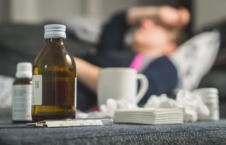 手を額に当ててt横たわる男性の手前に置かれた様々な種類の薬やマグカップ