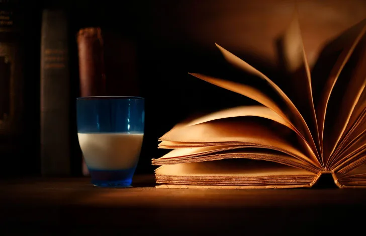 薄暗い机の上の本の横に置かれた水色のグラスに入った牛乳
