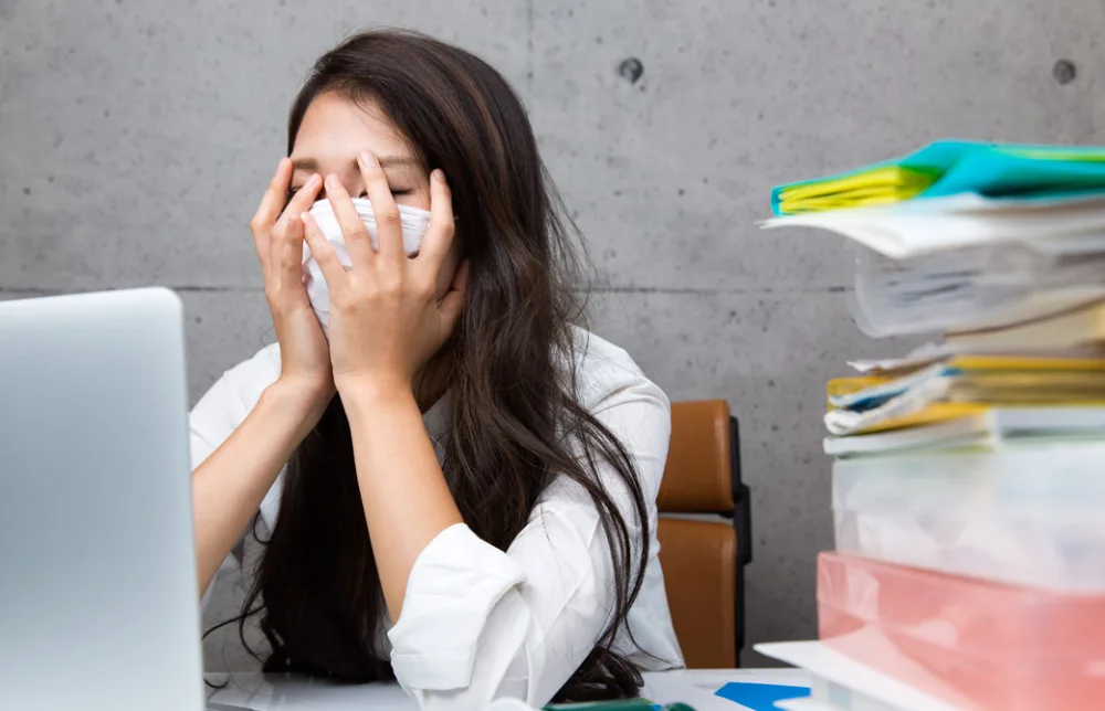 オフィスで、PCと山積みの書類を前に手で顔を覆う女性