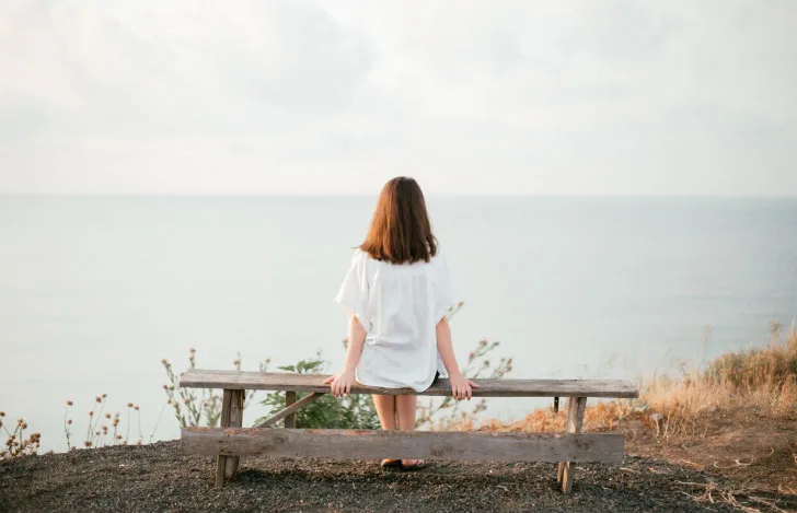 海辺のベンチに1人で座る女性の後ろ姿