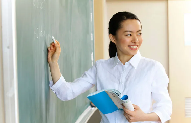 ノートを手に黒板に文字を書きながら笑顔で振り返る女性