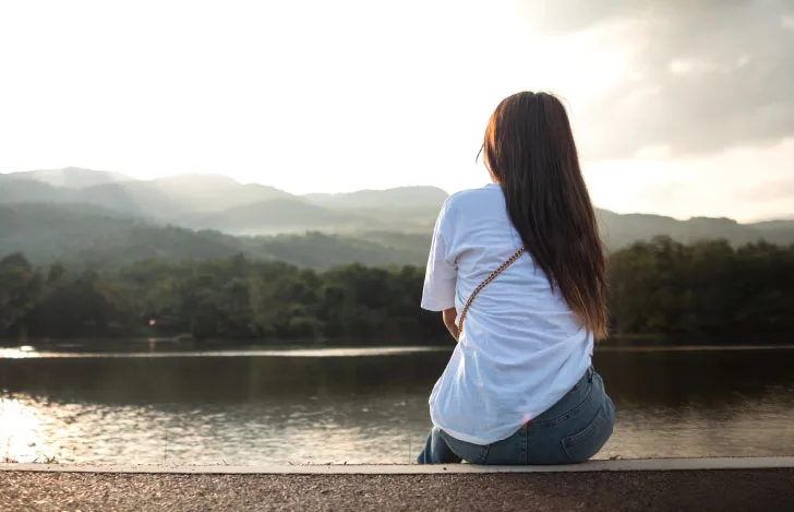 山と川を見ながら土手に座る女性の背中