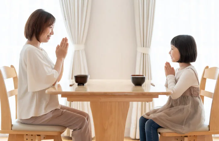 食卓で食事を前に向かい合って座り、手を合わせる親子